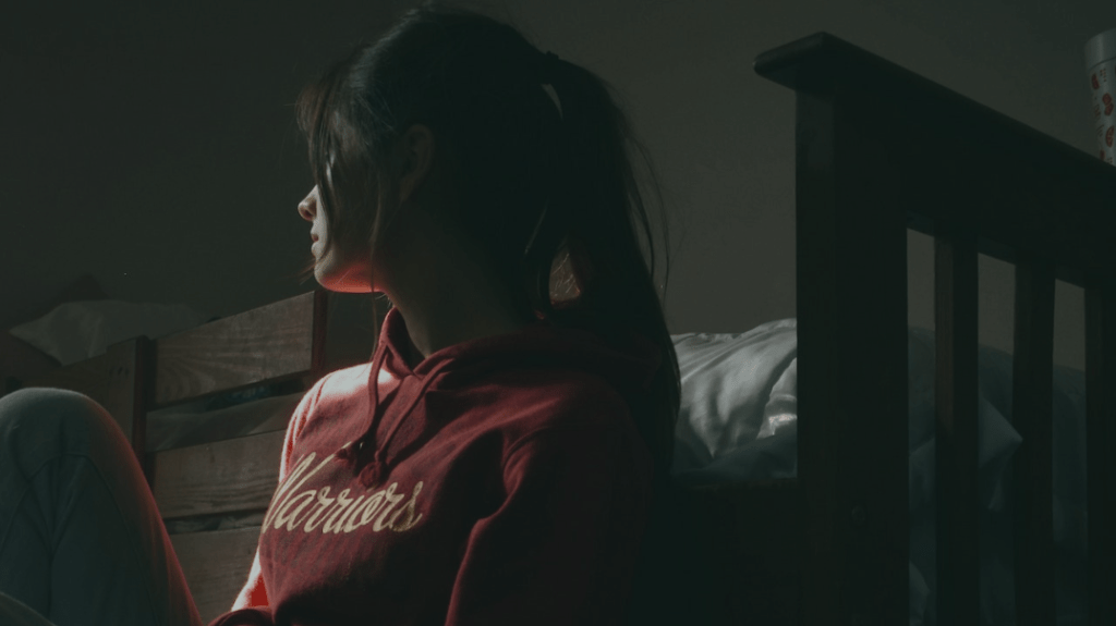 Woman with long ponytail, wearing a hoodie sits on the floor and looks away sadly.