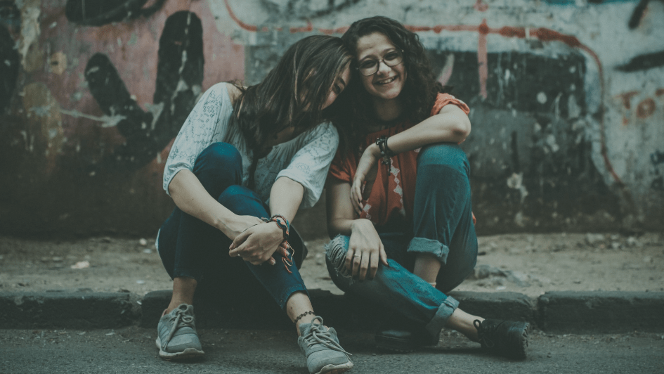 Two women sit together on a curb smiling at the camera.